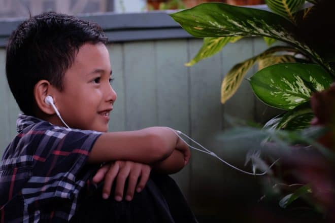 photo of a child sitting and listening to something on earbuds. There is a plant in front.