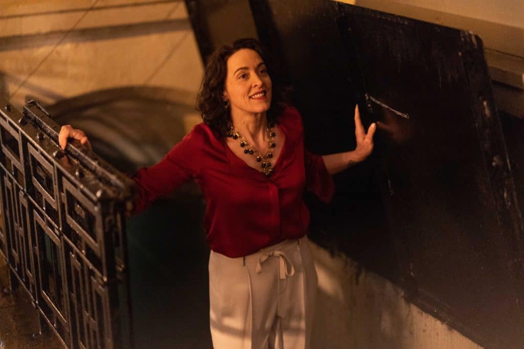 A middle-aged white woman with short, brown, curly hair in a dark red blouse, tan pants, and a gold and black statement necklace smiles as she emerges from a cellar. (credit: Facebook Watch)